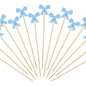 May include: A collection of light blue bow-shaped cupcake toppers on wooden sticks. The bows are a soft pastel blue, and the sticks are a natural wood color. These decorative toppers are arranged in a fan shape against a white background.