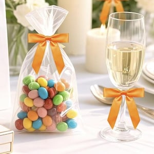 May include: A clear cellophane bag tied with an orange bow, filled with colorful candies. A champagne flute with an orange bow is next to the bag. White candles and flowers are in the background.