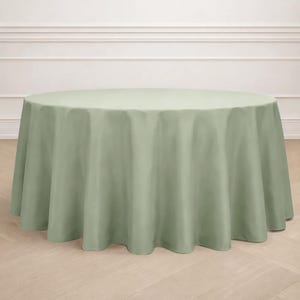 May include: A sage green round tablecloth. The tablecloth is draped over a round table, with the fabric falling in soft folds to the floor. The background features a light-colored wall with molding and a wooden floor.