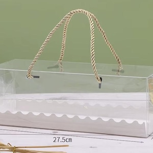May include: A clear rectangular box with a rope handle, ideal for packaging gifts or baked goods. The box is 27.5cm long, 11cm wide, and 9.5cm tall. The handle is made of twisted beige rope. The box has a white scalloped insert.