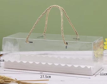 May include: A clear rectangular box with a rope handle, suitable for packaging gifts or baked goods. The box measures 27.5cm long, 11cm wide, and 9.5cm tall. The handle is made of twisted beige rope. The box has a white scalloped insert.