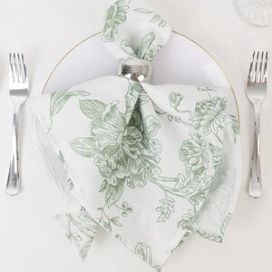 May include: A white cloth napkin with a green floral design, folded and held by a silver napkin ring, rests on a white plate. Two silver forks are on either side. The napkin's pattern features large flowers and leaves.