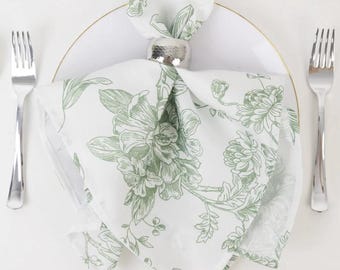 May include: A white napkin with a green floral pattern, held by a silver napkin ring, sits on a white plate. Two silver forks flank the plate. The napkin is patterned with large flowers and leaves, creating an elegant table setting.