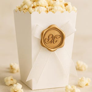 May include: A white popcorn box filled with popcorn, tied with a white ribbon and a gold seal. The seal has an ornate letter "H" in the center. Scattered popcorn kernels surround the box.