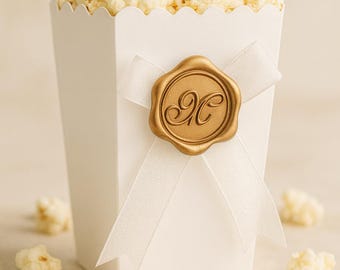 May include: A white popcorn box filled with popcorn, tied with a white ribbon and a gold seal. The seal features an ornate letter "H". Popcorn kernels are scattered around the box.