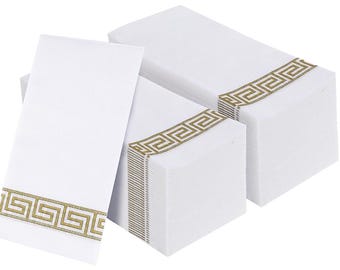 May include: A stack of white rectangular paper napkins featuring a gold-colored Greek key pattern border. The napkins are arranged in a group with one standing upright and two lying flat, showcasing the repeating geometric design on each piece.