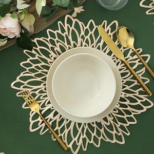 May include: A table setting with a cream-colored floral placemat, white plates, and gold cutlery. The arrangement includes a bowl, a dinner plate, a knife, a spoon, and a fork. The background is a dark green tablecloth, with a floral arrangement in the upper left corner.