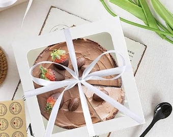May include: A chocolate cake in a white box, tied with a white ribbon. The cake is decorated with chocolate-covered strawberries and chocolate candies. The box has a clear window to view the cake.