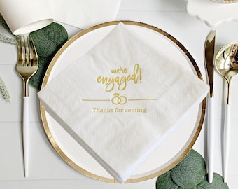 May include: White square paper napkins with gold text that reads "we're engaged!" and "Thanks for coming." The napkins are stacked on a white plate with a gold rim, surrounded by gold and white cutlery and greenery.