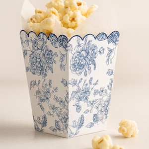May include: A white popcorn box with a blue floral design, filled with popcorn. The box has a scalloped edge and a classic toile pattern. Several pieces of popcorn are scattered around the box.