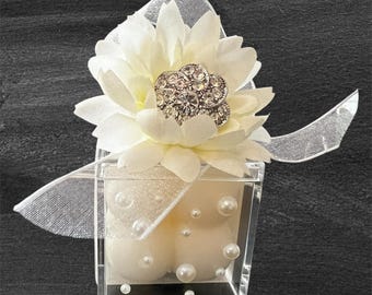 May include: A clear acrylic box containing two ivory candles and faux pearls, topped with a white fabric flower and a rhinestone embellishment. A sheer white ribbon is tied around the box, creating a decorative gift.