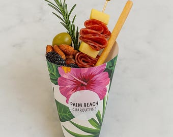 May include: A charcuterie cone filled with olives, almonds, blackberries, cheese, and salami. A rosemary sprig and breadstick are included. The cone is decorated with a pink hibiscus flower and the text "PALM BEACH CHARCUTERIE."