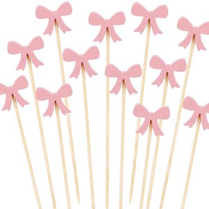 May include: A collection of pink bow-shaped decorative picks on wooden skewers. The bows are a soft pastel pink, and the skewers are a natural wood color. The picks are arranged against a white background.