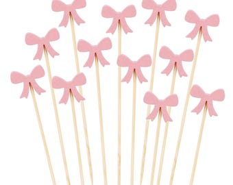May include: A selection of pink bow-shaped decorative picks on wooden skewers. The bows are a soft pastel pink, and the skewers are a natural wood colour. The picks are arranged against a white background.