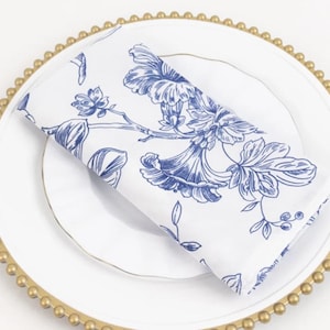 May include: A white folded napkin with a blue floral pattern rests on a white plate. The plate is on a gold beaded charger. The napkin features a detailed botanical design, adding a touch of elegance to the table setting.