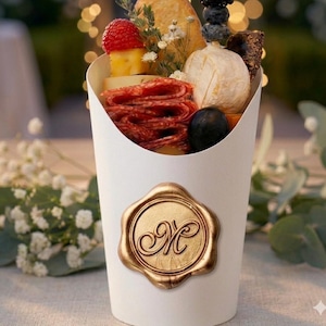 May include: A white paper cone filled with an assortment of gourmet snacks, including salami, cheese, fruit, and crackers. A gold-colored wax seal with the letter "M" is affixed to the front of the cone. The background is blurred with bokeh lights.