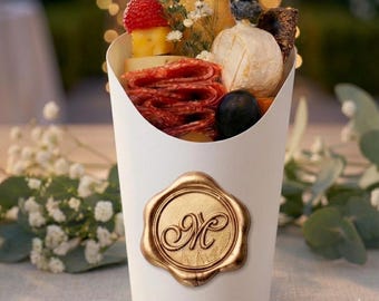 May include: A white paper cone filled with a selection of gourmet snacks, including salami, cheese, fruit, and crackers. A gold-coloured wax seal with the letter "M" is attached to the front. The background is blurred with bokeh lights.