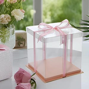 May include: A clear, square cake box with a pink base and a pink ribbon tied in a bow on top. The box is empty and sits on a white surface. Pink roses are in the foreground.