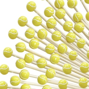 May include: A collection of yellow tennis ball-shaped decorative items on wooden skewers, arranged against a white background. The tennis balls have white curved stripes, and the skewers are made of light-colored wood.