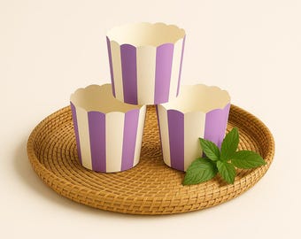 May include: Three paper cupcake liners with a vertical striped pattern in light purple and cream. The liners have a scalloped top edge and are arranged in a stack on a round, woven brown rattan tray, accompanied by a small sprig of green mint leaves.