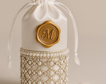 May include: A white satin favor bag with a gold seal bearing the letter "M". The lower section is decorated with a pearl and rhinestone design. Several white almond sweets are placed nearby. A decorative item.