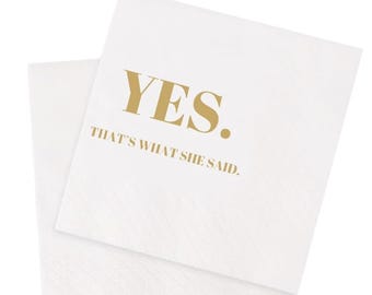May include: Two white paper napkins with a textured surface. The top napkin has the word "YES." printed in gold, with the phrase "THAT'S WHAT SHE SAID." below it, also in gold. The napkins are likely for a party or event.