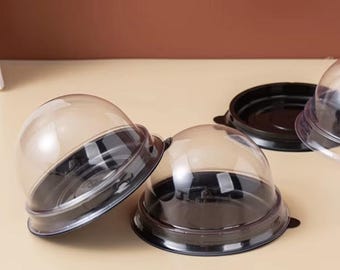 May include: Clear plastic dome-shaped containers with black bases. The containers are designed to hold baked goods or other small items. The image shows several containers, some with lids attached and others without.