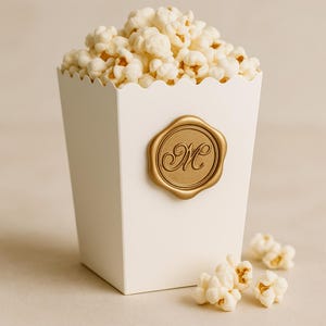 May include: A white popcorn box filled with fluffy popcorn. The box has a scalloped top and a gold seal with the letter "M". Several pieces of popcorn are scattered around the base of the box. The overall theme is a snack or party favor.