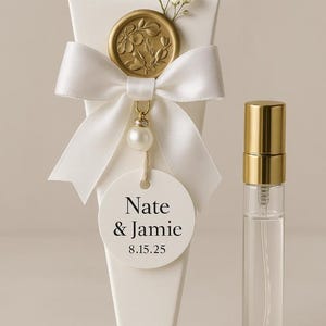 May include: A white favor box with a gold floral seal, white ribbon, and a tag that reads "Nate & Jamie 8.15.25." A small perfume bottle with a gold cap is next to the box. The box is decorated with a sprig of white flowers.