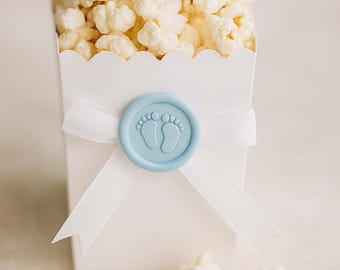 May include: A white popcorn box filled with fluffy popcorn, tied with a white ribbon and sealed with a light blue wax seal featuring baby footprints. A few pieces of popcorn are scattered around the box.