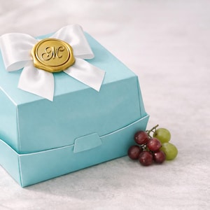 May include: A light blue gift box with a white ribbon and a gold seal. The seal has the letter "M" in the center. A small bunch of red and green grapes sits next to the box.