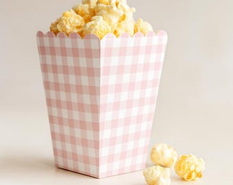 May include: A pink and white gingham popcorn box filled with caramel popcorn. Three pieces of popcorn are scattered around the box. The box has a scalloped top edge and a trapezoid shape.