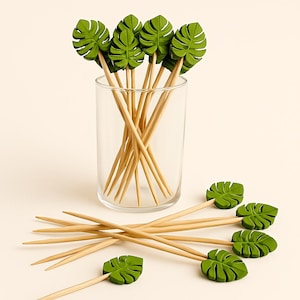 May include: A collection of decorative cocktail picks with green leaf-shaped toppers. The picks are made of wood and are arranged in a clear glass and scattered around it. The leaf toppers are a vibrant green color.