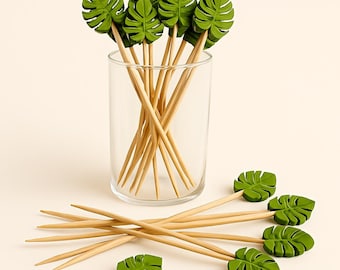 May include: A collection of decorative cocktail picks with green leaf-shaped toppers. The picks are made of wood and are arranged in a clear glass and scattered around it. The leaf toppers are a vibrant green colour.