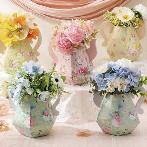 May include: Five hexagonal paper vases with floral patterns, each featuring two side handles and filled with artificial flowers in yellow, orange, pink, and blue. The vases are arranged in two rows, displaying various botanical prints and pastel colors.