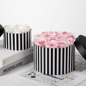 May include: Two round gift boxes with black and white vertical stripes. The smaller box contains white roses, while the larger box is filled with light pink roses. Each box has a separate black lid placed to the side of the container.