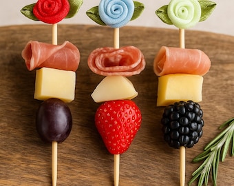 May include: Three decorative skewers with food items, including cheese, grapes, salami, strawberries, and blackberries. Each skewer is topped with a fabric flower in red, blue, and green. The skewers are arranged on a wooden cutting board.