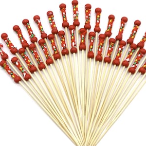 May include: A collection of decorative cocktail skewers. Each skewer has a natural wood shaft and a red, decorative top with colorful accents. The skewers are arranged in a fan shape against a white background.