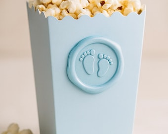 May include: A pale blue popcorn container filled with popcorn. The container has a scalloped edge and a circular embossed design of baby footprints. Popcorn pieces are scattered around the base of the container.