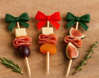 May include: Three decorative appetizer skewers with a green, and red bow on top. Each skewer features a different combination of food items, including cheese, salami, grapes, apricots, and figs. The skewers are arranged on a wooden surface.