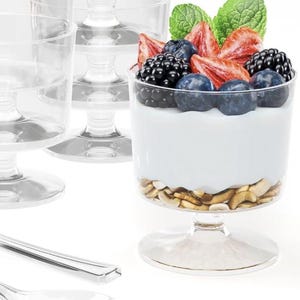 May include: Clear plastic dessert cups with a pedestal base. One cup is filled with yogurt, granola, and topped with fresh strawberries, blueberries, blackberries, and mint leaves. A silver spoon is in the foreground.