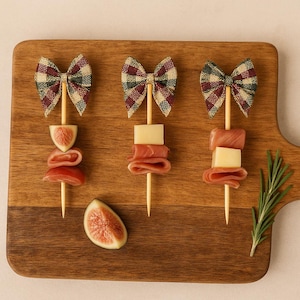May include: Three appetizer skewers with bow-tie toppers, featuring prosciutto, cheese, and fig slices. The skewers are arranged on a wooden cutting board with a sprig of rosemary and a half fig.