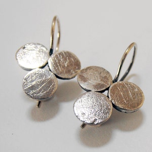 May include: A pair of silver earrings featuring a cluster of three textured, circular discs. The earrings have a simple hook design for pierced ears. The discs have a scratched surface, giving them a unique, handcrafted appearance.