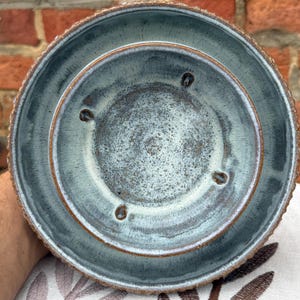 May include: Overhead view of a ceramic planter with a textured, ridged rim and a two-tiered design. The planter has a mottled blue-green glaze with brown accents. Three drainage holes are visible in the inner tier.