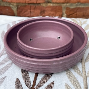 May include: A dusty rose ceramic planter set. The set includes a large, shallow dish and a smaller, circular pot with two drainage holes. The pieces have a ridged texture and a matte finish. The set is designed for indoor or outdoor use.