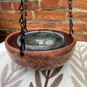 May include: A hanging planter with a speckled coral-colored outer bowl and a smaller, speckled gray inner bowl. The bowls are suspended by a black chain against a brick wall background. The planter sits on a patterned fabric.