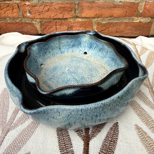 May include: Two ceramic bowls in a nested arrangement. The bowls have a wavy rim and a speckled blue glaze with dark blue and brown accents. The bowls are sitting on a patterned white and tan surface.