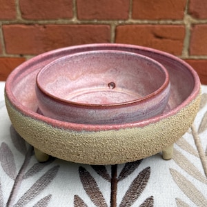 Pode incluir: Um conjunto de duas taças de cerâmica encaixáveis com um exterior texturizado em bege areia e um interior esmaltado liso em tons de rosa. A taça maior tem três pés pequenos, enquanto a taça menor está colocada no seu interior.