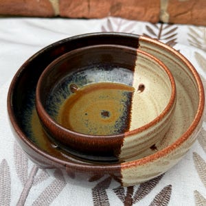 May include: A set of three handcrafted ceramic bowls. The bowls are nested, with a dark brown exterior and a two-tone interior of tan and brown. The bowls have a glossy finish and are likely used for serving or display.