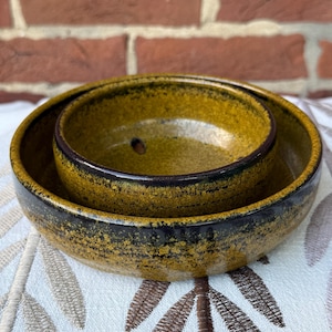 May include: Two ceramic bowls, one nested inside the other. The bowls have a speckled, mustard-yellow glaze with a dark brown rim. The outer bowl is larger, and the inner bowl has a small hole in the center.